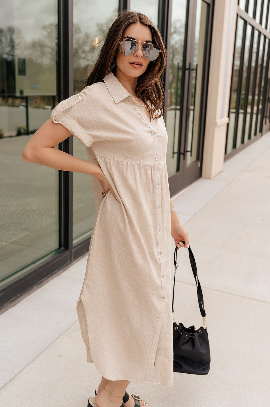 Full body side view of model wearing the Daphne Oatmeal Button-Up Midi Dress which features beige fabric, a button-up front with pearlescent buttons, a collared neckline, short sleeves with cuffs and button tabs, and a rounded midi hemline.