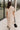 Full body back view of model wearing the Daphne Oatmeal Button-Up Midi Dress which features beige fabric, a button-up front with pearlescent buttons, a collared neckline, short sleeves with cuffs and button tabs, and a rounded midi hemline.