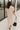 Full body back view of model wearing the Daphne Oatmeal Button-Up Midi Dress which features beige fabric, a button-up front with pearlescent buttons, a collared neckline, short sleeves with cuffs and button tabs, and a rounded midi hemline.