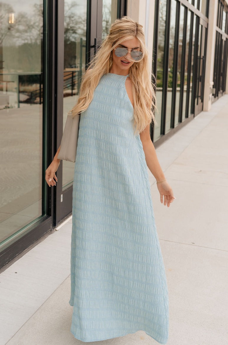 Full body view of model wearing the Gillie Light Blue Halter Midi Dress which features light blue and yellow knit textured fabric, light blue lining, small gingham print design, midi length, halter neckline, adjustable straps, sleeveless and open back.