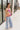 Full body side view of model wearing the Bianca Satin Floral Tank Top that has sage green satin fabric with a green, purple, pink, yellow, and red floral print and a halter neckline with 3 back button closures. Top is shown untucked.