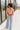 Full body back view of model wearing the Bianca Satin Floral Tank Top that has sage green satin fabric with a green, purple, pink, yellow, and red floral print and a halter neckline with 3 back button closures. 