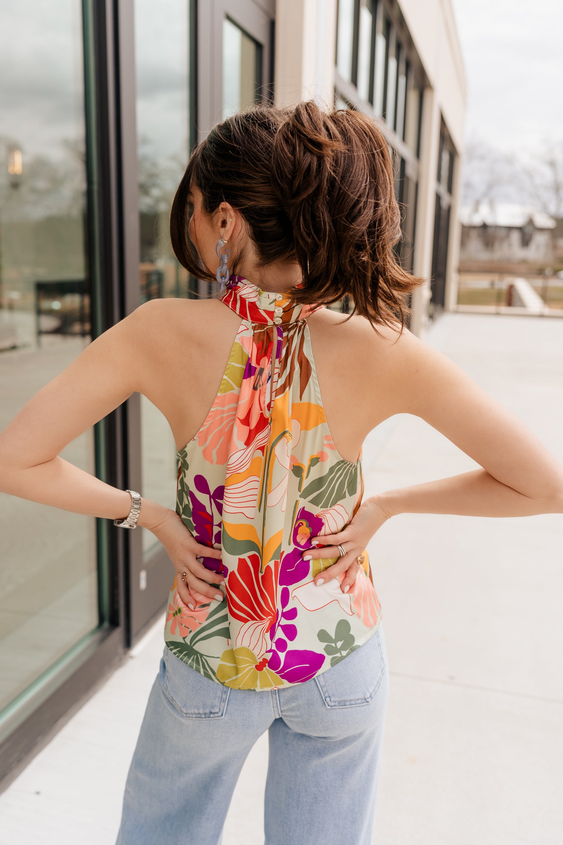 Back view of model wearing the Bianca Satin Floral Tank Top that has sage green satin fabric with a green, purple, pink, yellow, and red floral print and a halter neckline with 3 back button closures. Top is shown untucked.
