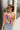 Front view of model wearing the Bianca Satin Floral Tank Top that has sage green satin fabric with a green, purple, pink, yellow, and red floral print and a halter neckline with 3 back button closures. Top is shown tucked into jeans.