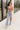 Full body front view of model wearing the Bianca Satin Floral Tank Top that has sage green satin fabric with a green, purple, pink, yellow, and red floral print and a halter neckline with 3 back button closures. Top is shown tucked into jeans.