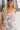 Close upper front view of model wearing the Gwen White Floral Mini Dress that features white fabric with a peach, brown, green, blue, and purple floral print, adjustable spaghetti straps, a smocked back with a zipper and hook closure, a flared skirt with a structured hemline, and white lining.