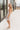 Full body side view of model wearing the Gwen White Floral Mini Dress that features white fabric with a peach, brown, green, blue, and purple floral print, adjustable spaghetti straps, a smocked back with a zipper and hook closure, a flared skirt with a structured hemline, and white lining.