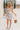 Full body front view of model wearing the Gwen White Floral Mini Dress that features white fabric with a peach, brown, green, blue, and purple floral print, adjustable spaghetti straps, a smocked back with a zipper and hook closure, a flared skirt with a structured hemline, and white lining.