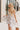 front view of model wearing the Gwen White Floral Mini Dress that features white fabric with a peach, brown, green, blue, and purple floral print, adjustable spaghetti straps, a smocked back with a zipper and hook closure, a flared skirt with a structured hemline, and white lining.