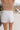 The girl model is wearing white denim shorts with a frayed hem. This is a back view of her wearing the shorts with a tan tank top.