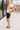 Full body side view of female model wearing the Dana Black Frayed Hem Denim Shorts that feature black distressed denim, frayed hems, and pockets. Paired with white top.