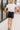 Full body back view of female model wearing the Dana Black Frayed Hem Denim Shorts that feature black distressed denim, frayed hems, and pockets. Paired with white top.