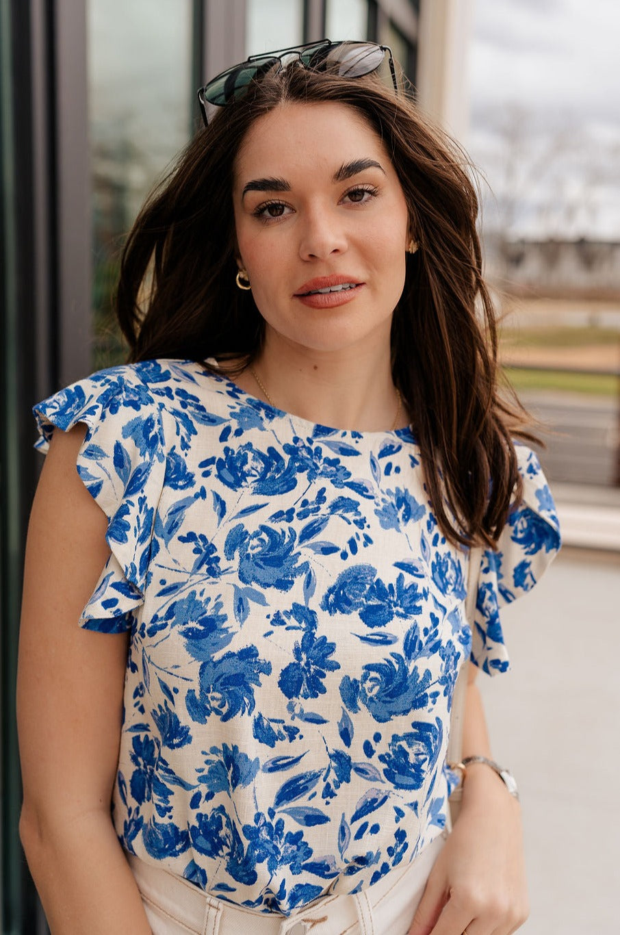 Close up view of model wearing the Carmen Blue & Cream Floral Top which features blue and cream knit fabric, floral pattern, round neckline, ruffle short sleeves and keyhole back with button closure.