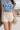 Close-up front view of female brunette model wearing the Mckenzie Off White Denim Shorts that feature off white denim, contrast stitching, pockets, and a high rise. Styled with blue and white floral top.