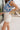 Side view of female brunette model wearing the Mckenzie Off White Denim Shorts that feature off white denim, contrast stitching, pockets, and a high rise. Styled with blue and white floral top.
