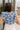 Back view of model wearing the Carmen Blue & Cream Floral Top which features blue and cream knit fabric, floral pattern, round neckline, ruffle short sleeves and keyhole back with button closure.