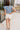 Full body back view of female brunette model wearing the Mckenzie Off White Denim Shorts that feature off white denim, contrast stitching, pockets, and a high rise. Styled with blue and white floral top.