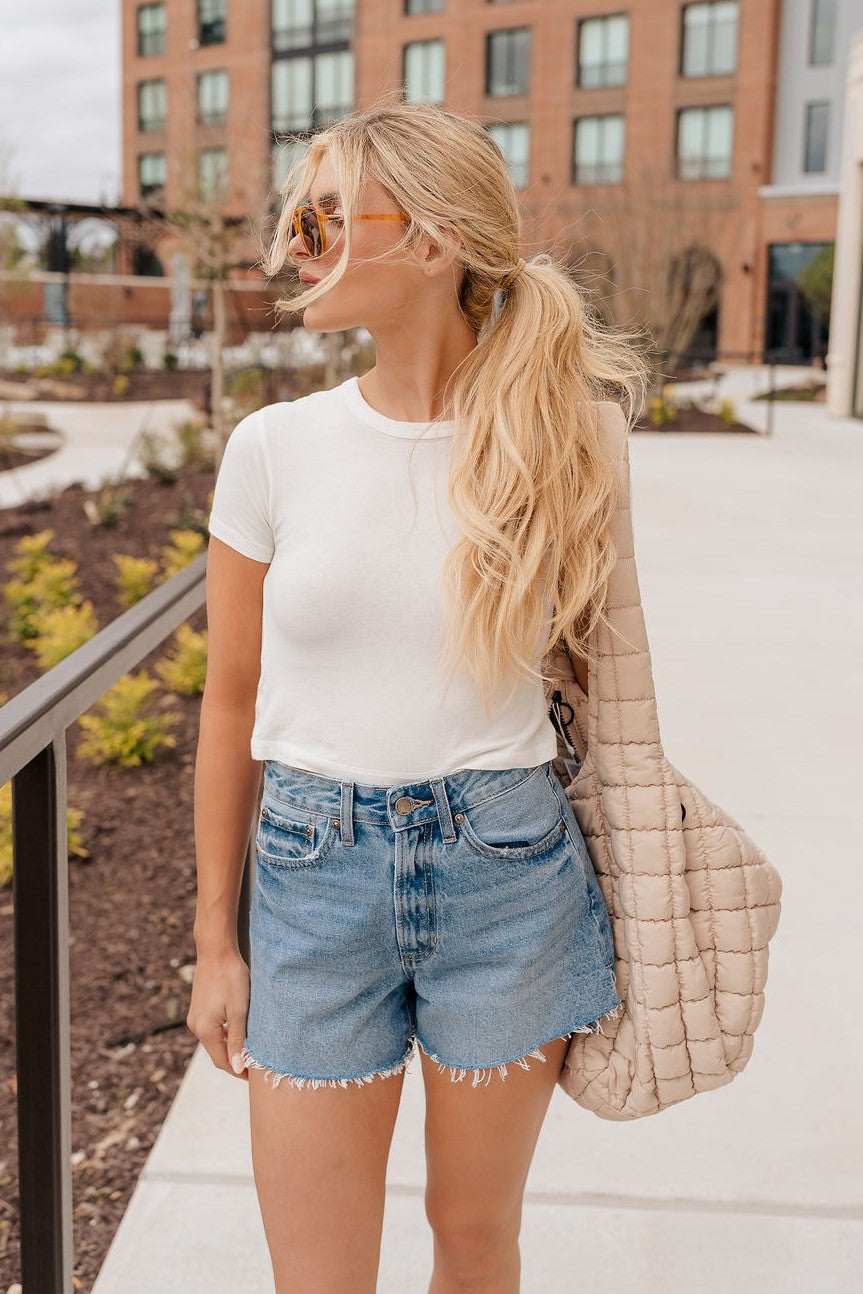 Front view of female blonde model wearing the medium wash color of the Hayden Frayed Hem Denim Shorts, paired with a white cropped t-shirt.