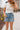 Front view of female blonde model wearing the medium wash color of the Hayden Frayed Hem Denim Shorts, paired with a white cropped t-shirt.