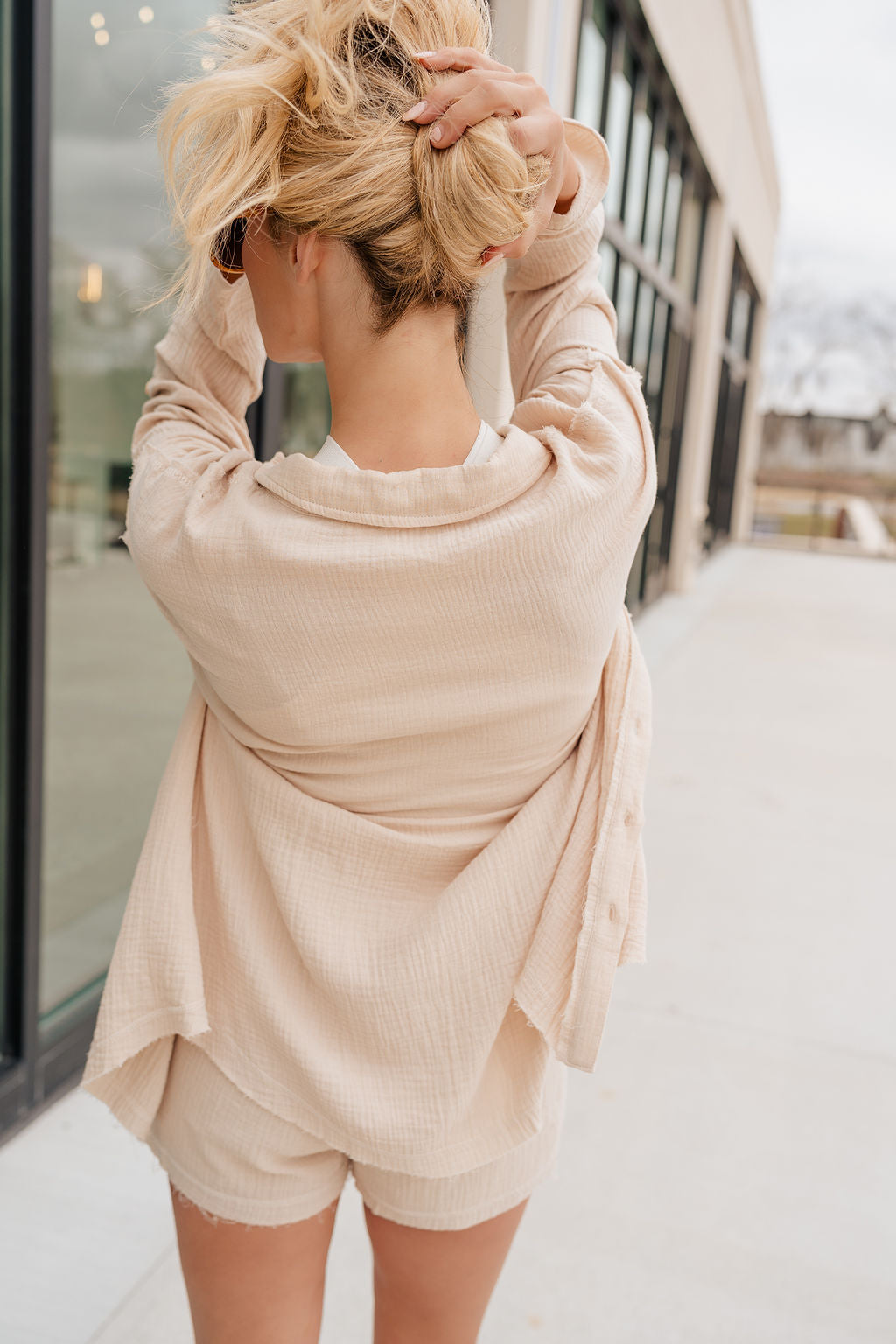 Back view of model wearing the Cassidy Beige Gauze Long Sleeve Top that features beige gauze fabric, a button-up front with beige button closures, a collared neckline, a left front chest pocket, drop shoulder long sleeves, and frayed hem details. Top is worn over white tank.