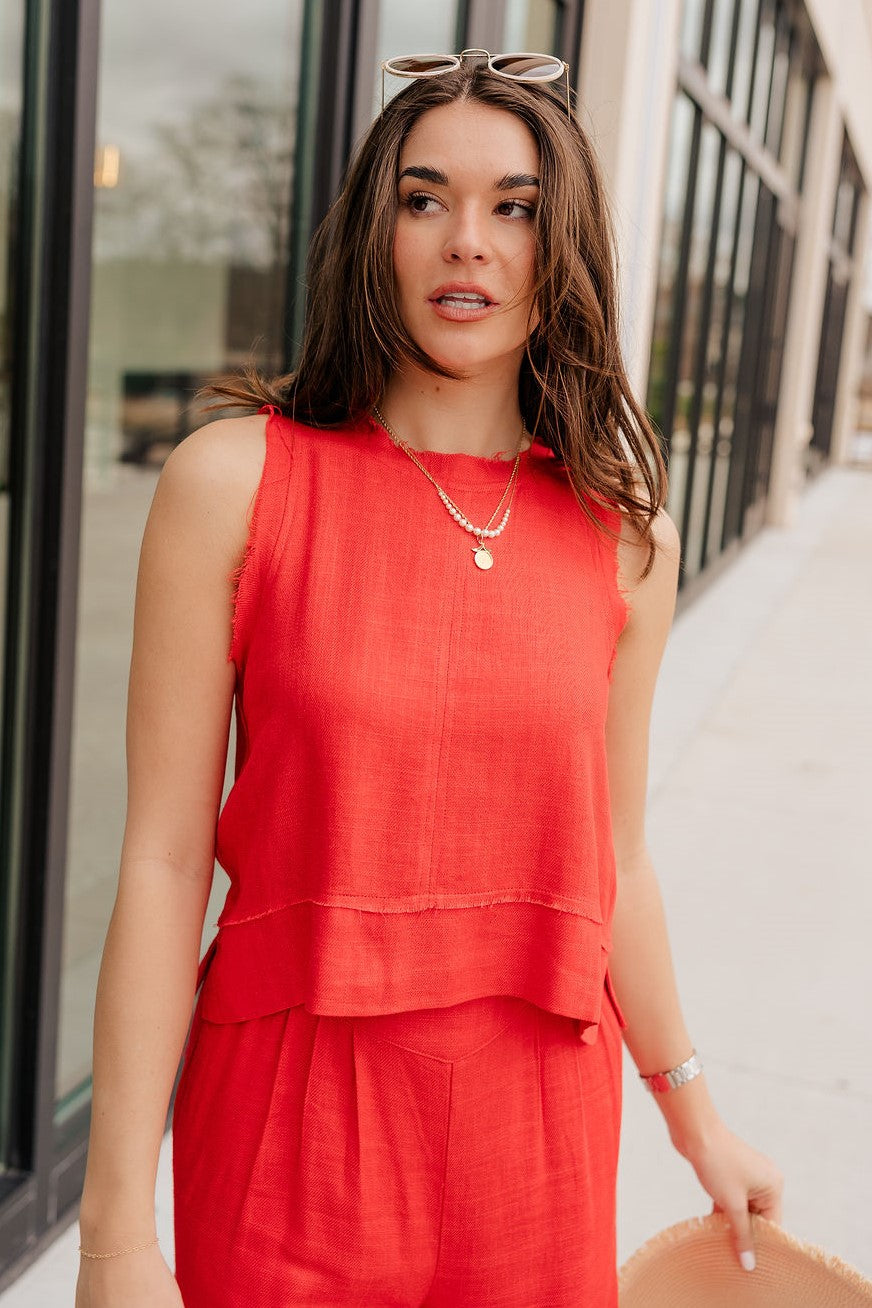 Front view of model wearing the Vada Red Tank Top that features red woven fabric with raw frayed hems, a round neckline, a sleeveless body, small side slits, and a keyhole back with a button closure.