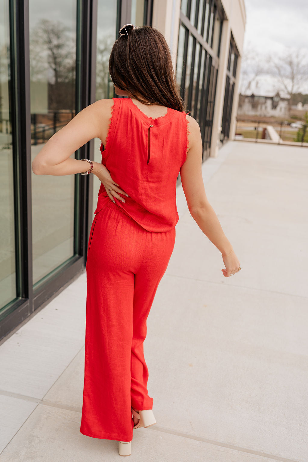 Full body back view of model wearing the Vada Red Tank Top that features red woven fabric with raw frayed hems, a round neckline, a sleeveless body, small side slits, and a keyhole back with a button closure.