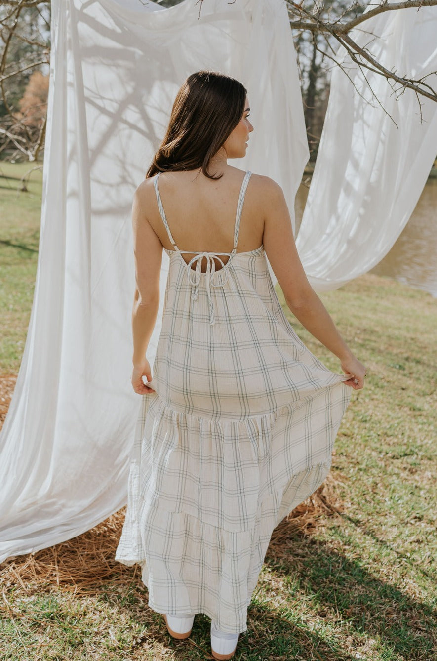 Full body back view of female model wearing the Milana Ivory & Blue Plaid Maxi Dress that has ivory fabric with blue plaid, spaghetti straps, tiered skirt, and ties in the back.