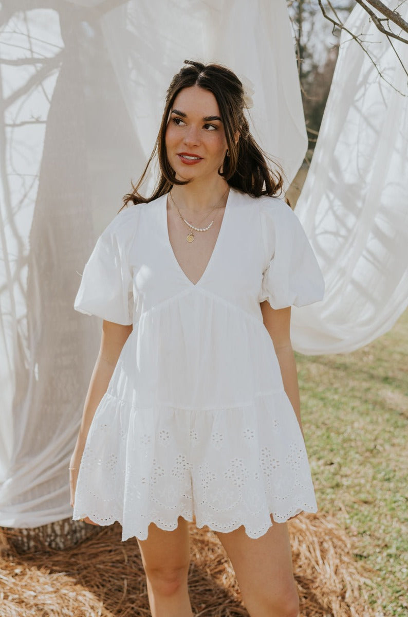 Front view of female model wearing the Madeline White Eyelet Romper that has white fabric with eyelet details, short puff sleeves, a vneck, and a keyhole back with a tie.