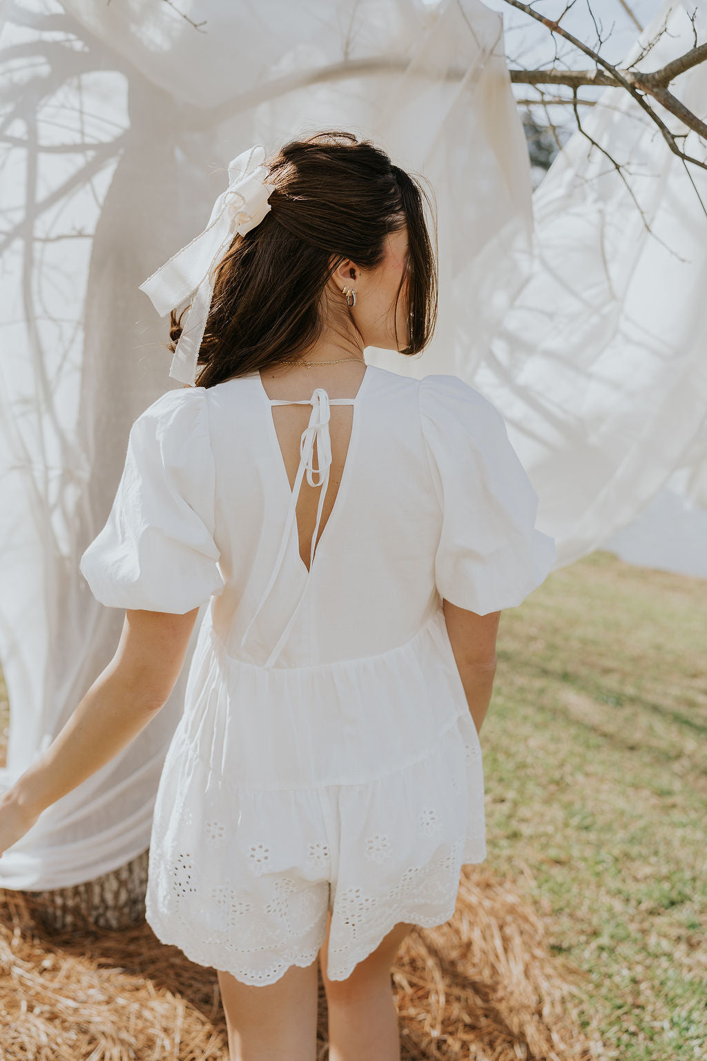 Back view of female model wearing the Madeline White Eyelet Romper that has white fabric with eyelet details, short puff sleeves, a vneck, and a keyhole back with a tie.