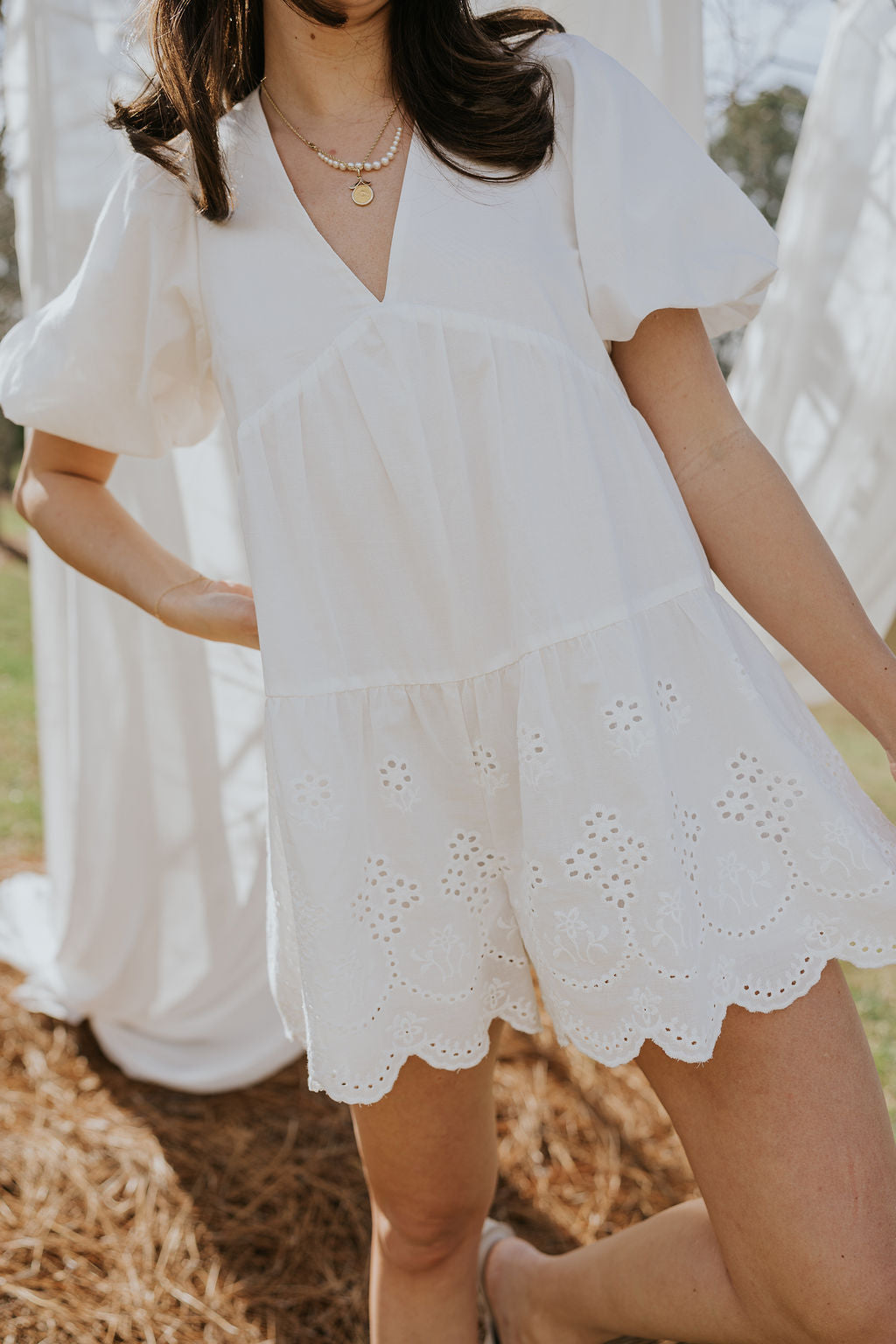 Close front view of female model wearing the Madeline White Eyelet Romper that has white fabric with eyelet details, short puff sleeves, a vneck, and a keyhole back with a tie.