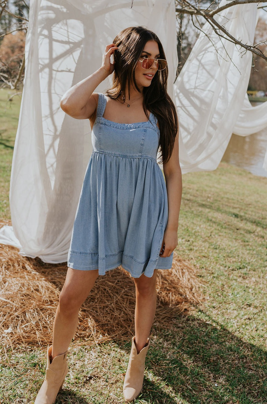 Full body front view of female model wearing the Tinsley Chambray Sleeveless Mini Dress that has blue chambray fabric, thick straps that have elastic in the back, and side pockets.