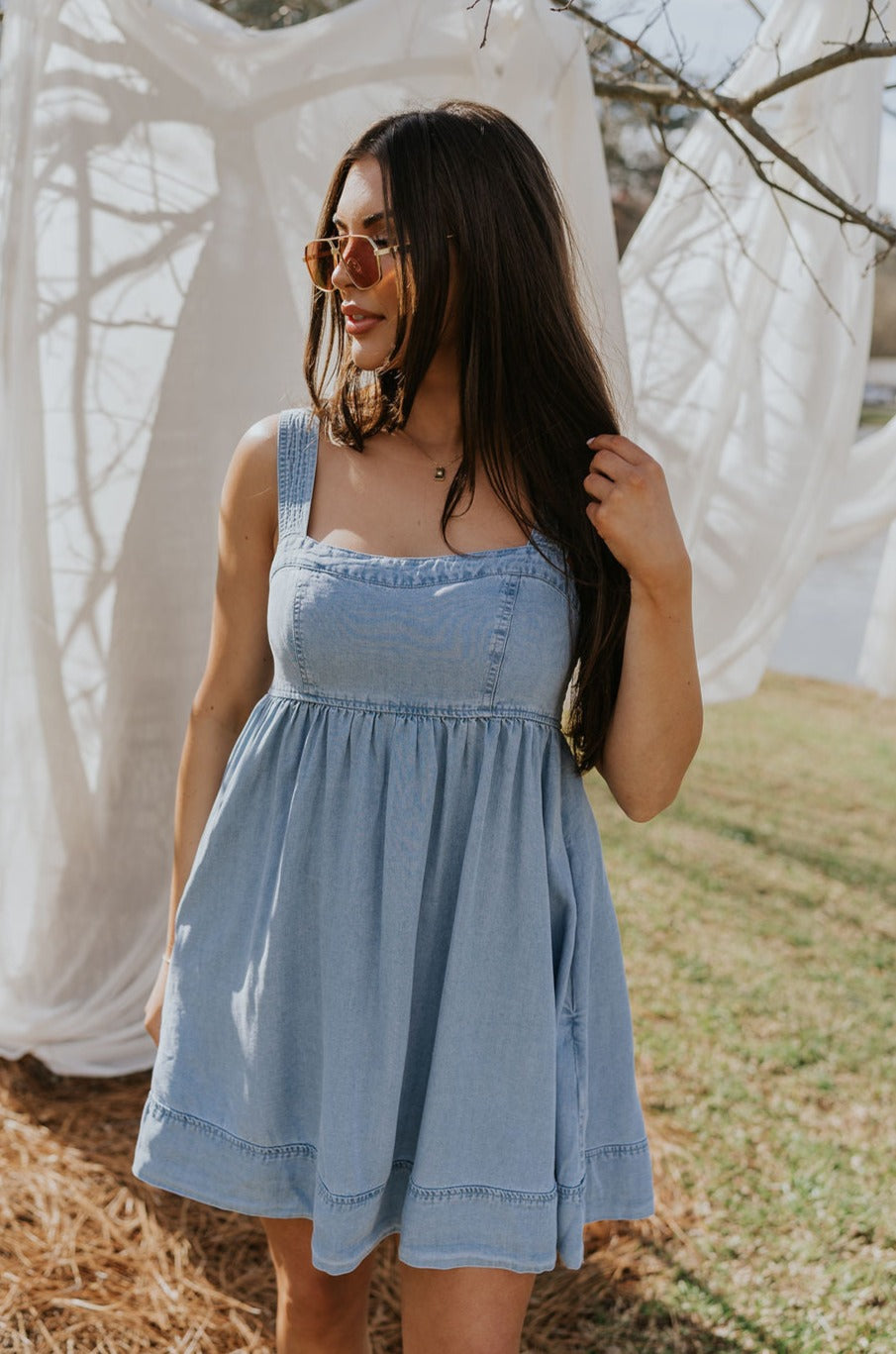Front view of female model wearing the Tinsley Chambray Sleeveless Mini Dress that has blue chambray fabric, thick straps that have elastic in the back, and side pockets.