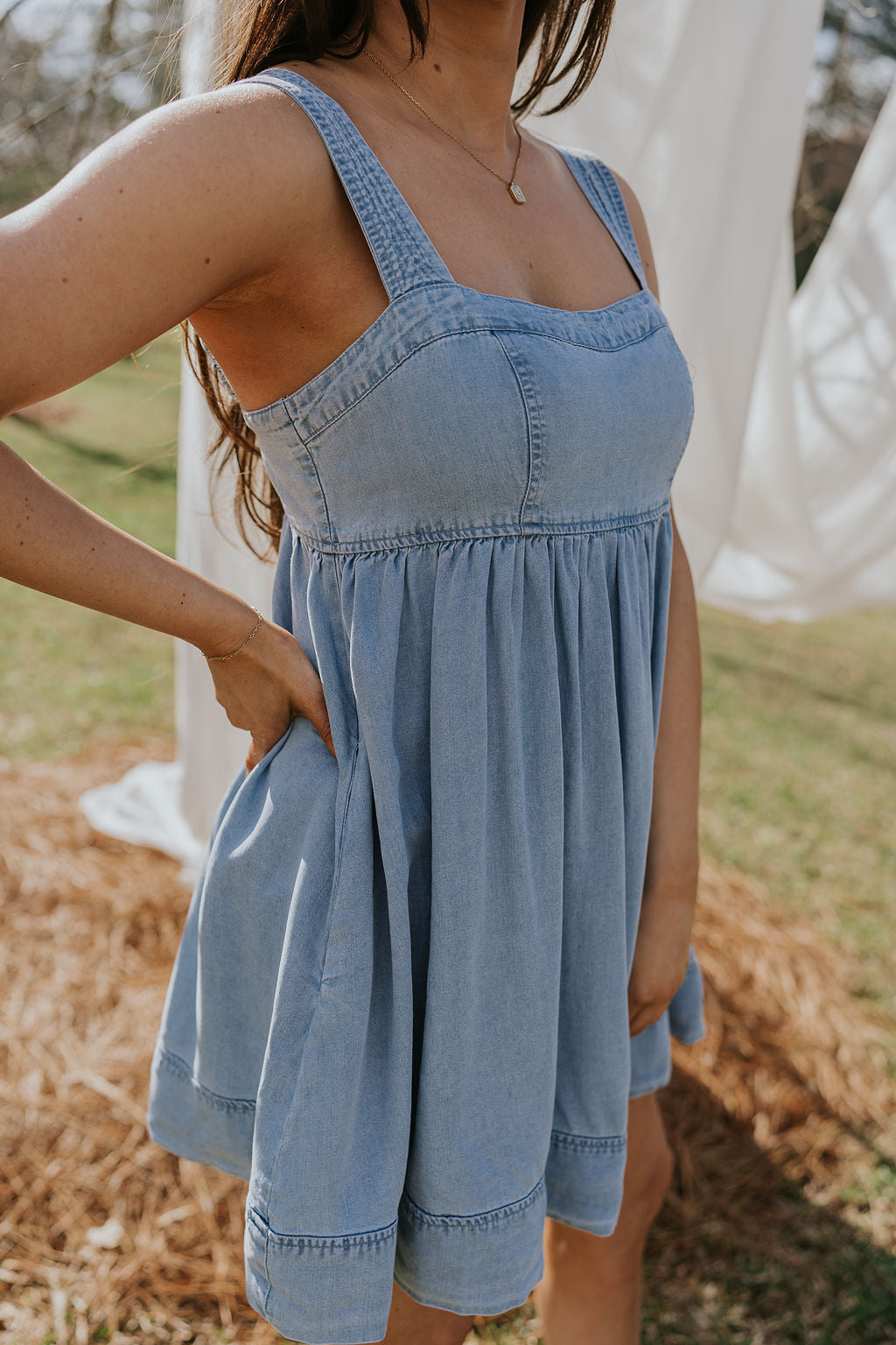 Side view of female model wearing the Tinsley Chambray Sleeveless Mini Dress that has blue chambray fabric, thick straps that have elastic in the back, and side pockets.