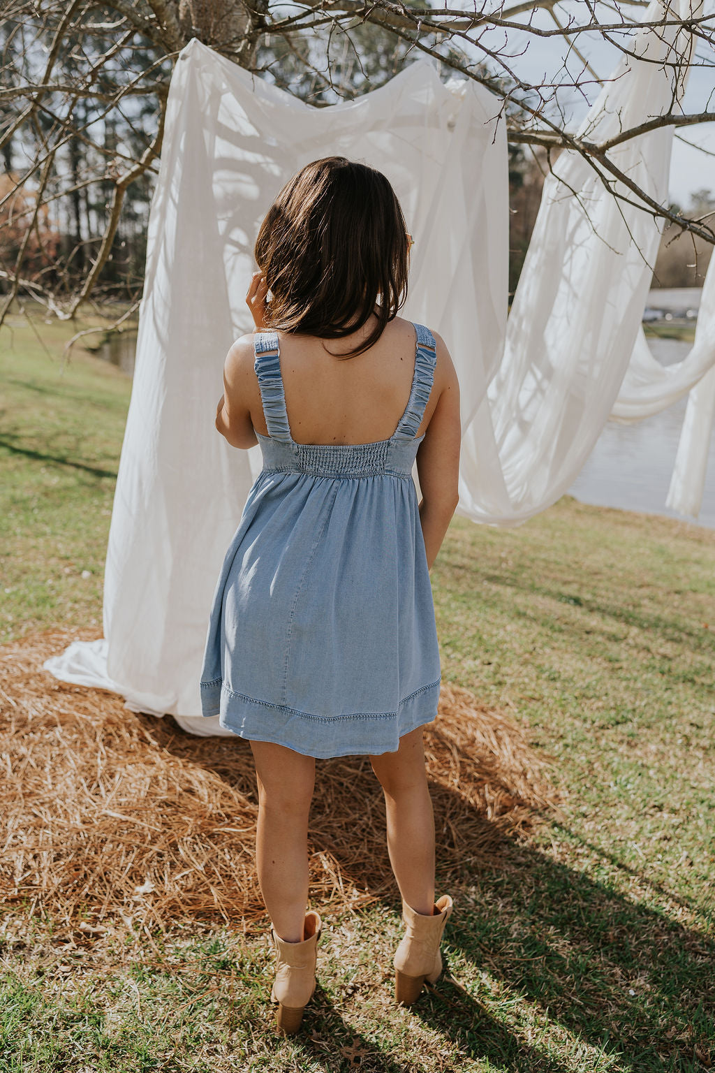 Full body back view of female model wearing the Tinsley Chambray Sleeveless Mini Dress that has blue chambray fabric, thick straps that have elastic in the back, and side pockets.
