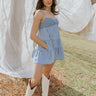 Full body front view of model wearing the Carla Chambray Sleeveless Mini Dress that has light blue fabric, tiered skirt, a smocked sweetheart neck and spaghetti straps. Worn with white boots.