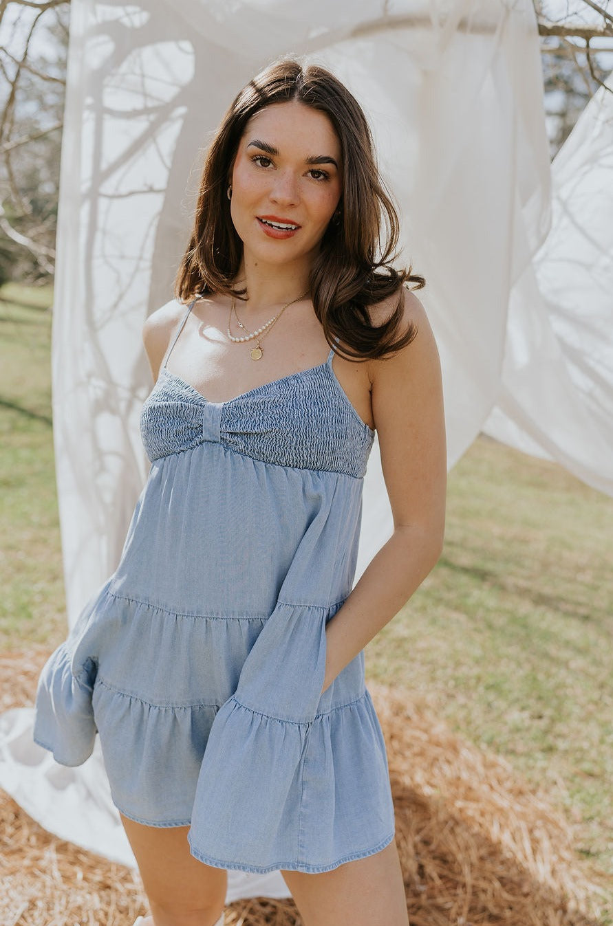 Front view of model wearing the Carla Chambray Sleeveless Mini Dress that has light blue fabric, tiered skirt, a smocked sweetheart neck and spaghetti straps. Worn with white boots.