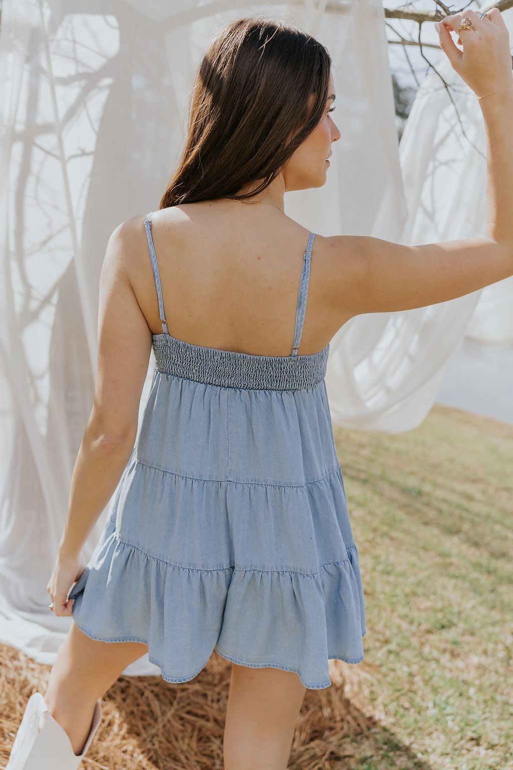 Back view of model wearing the Carla Chambray Sleeveless Mini Dress that has light blue fabric, tiered skirt, a smocked sweetheart neck and spaghetti straps. Worn with white boots.