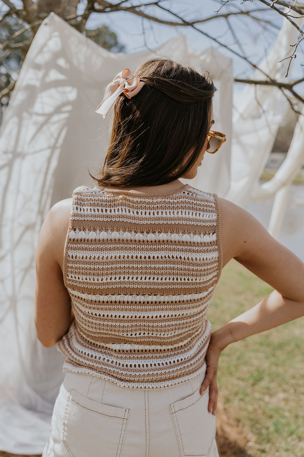 Back view of female model wearing the Mallory Taupe & Cream Crochet Tank that has taupe and cream crochet fabric, an open front with a tie, and a sleeveless body. Worn with cream denim shorts.