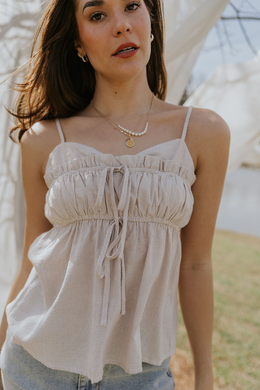 Close-up front view of female model wearing the Billie White & Taupe Striped Tank that has thin vertical stripes, front tie details, ruffle trim, and spaghetti straps