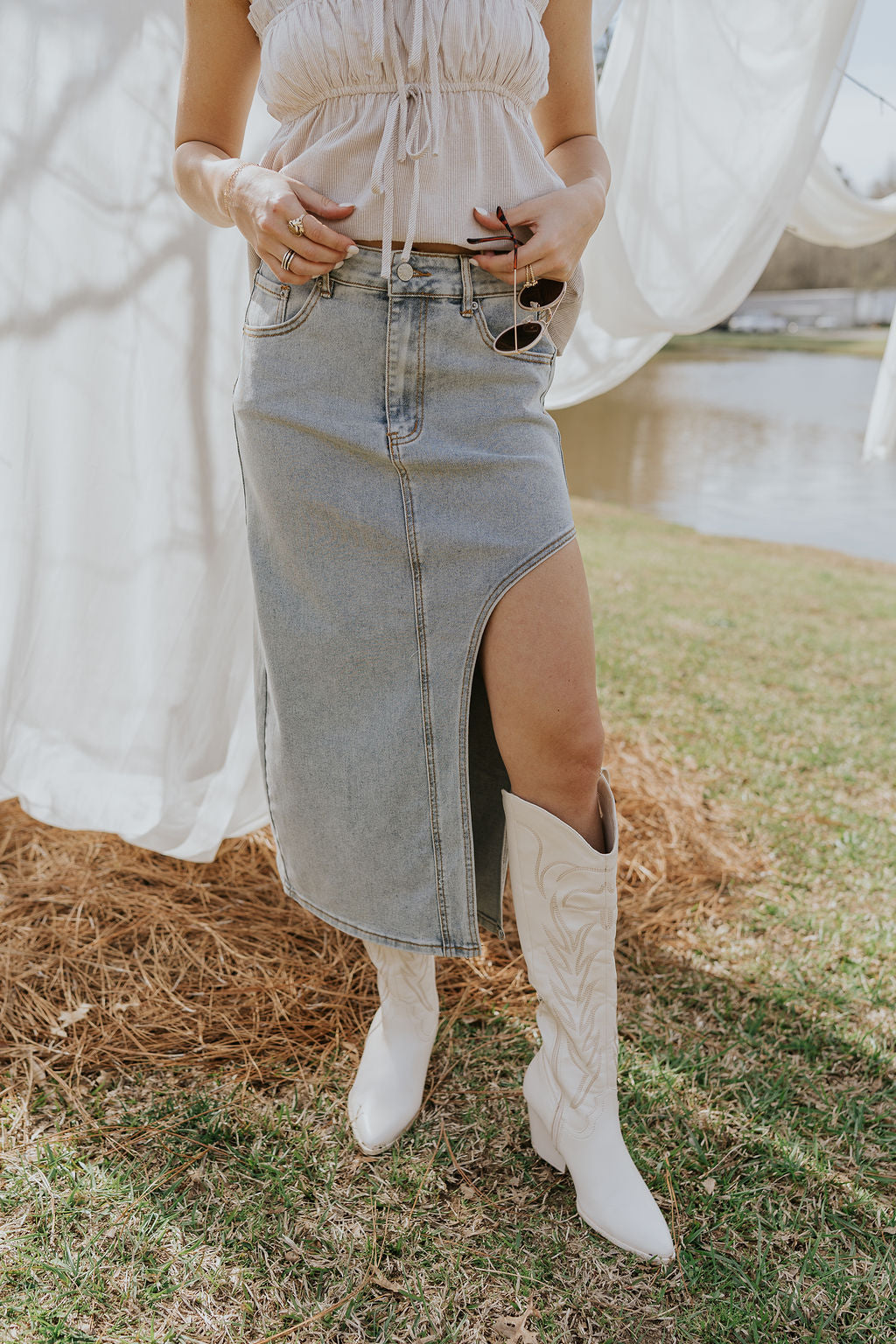 Front view of female model wearing the Lexie Asymmetric Denim Midi Skirt that has light wash denim, a slit cutout, midi-length hem, pockets, and belt loops. Worn with taupe tank top.