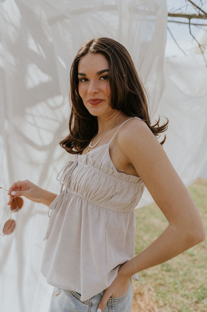 Side view of female model wearing the Billie White & Taupe Striped Tank that has thin vertical stripes, front tie details, ruffle trim, and spaghetti straps