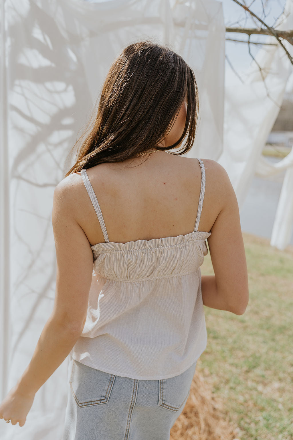 Back view of female model wearing the Billie White & Taupe Striped Tank that has thin vertical stripes, front tie details, ruffle trim, and spaghetti straps
