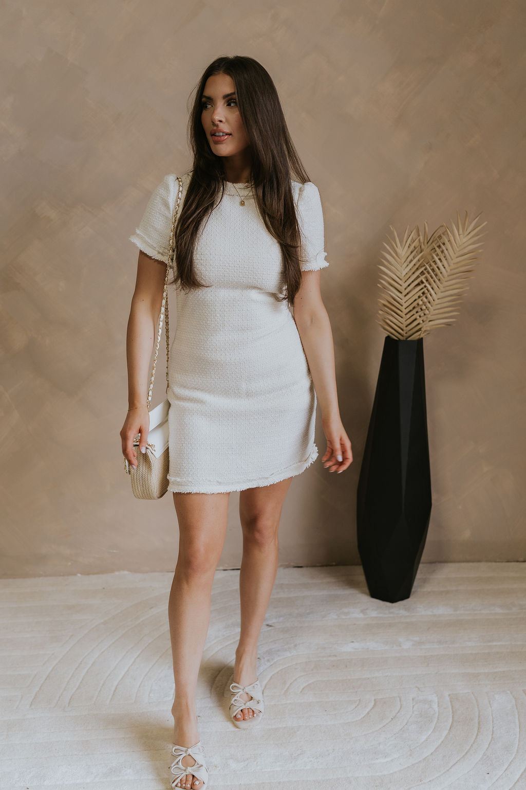 Full body front view of 5'5" female model wearing the Demi Tweed Short Sleeve Mini Dress in Off White that has off white tweed fabric, short sleeves with puff shoulders, a high round neck, and back zipper.
