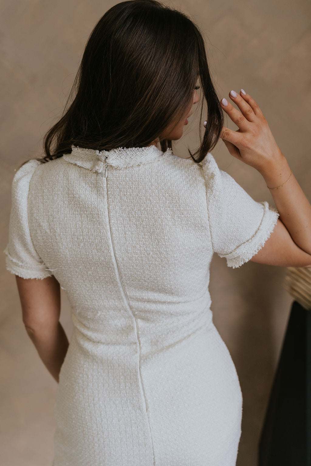 Upper body back view of 5'5" female model wearing the Demi Tweed Short Sleeve Mini Dress in Off White that has off white tweed fabric, short sleeves with puff shoulders, a high round neck, and back zipper.
