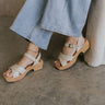 Left side view of female model wearing the Dasha Sandal in Off White which features off white upper leather fabric, kork platform sole, block heel, criss-cross strap and gold adjustable buckle closure