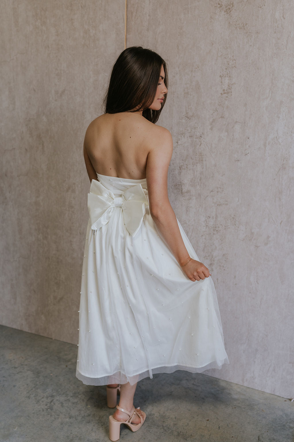 Full body back view of female model wearing the Rebecca Off White Pearl Bow Midi Dress which featuresCream Sheer Fabric, Midi Length, Cream Lining, Strapless, Pearl Details and Bow Back Detail