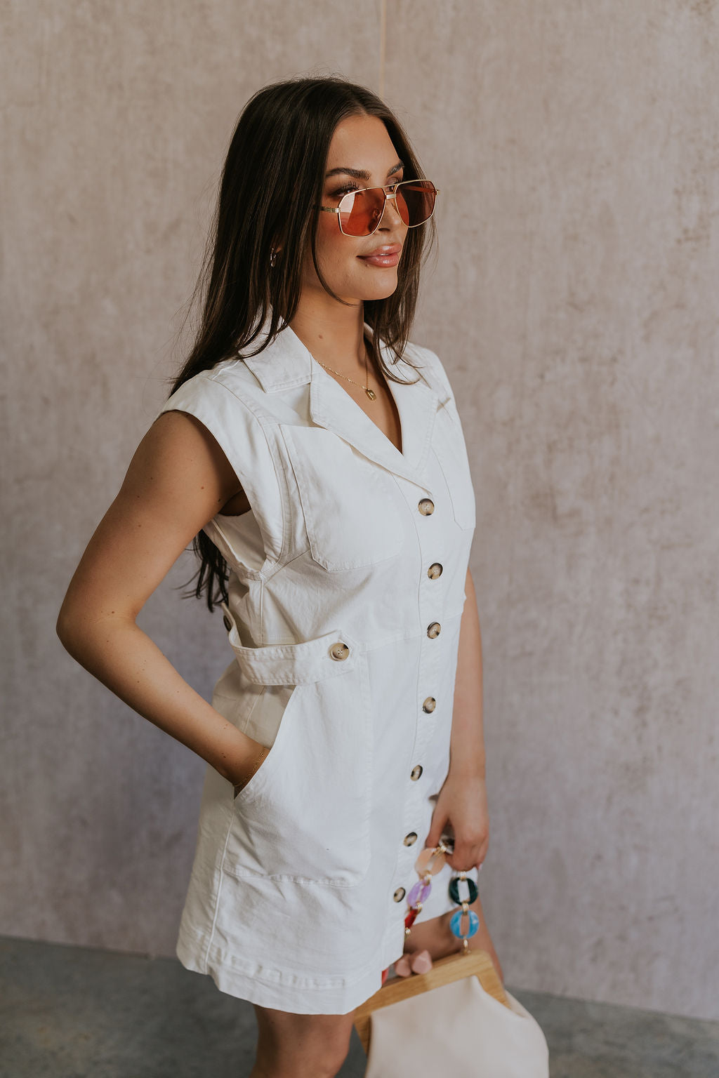 Side view of female model wearing the Jessica Off White Tortoise Button-Up Mini Dress which features Off White Fabric, Tortoise Button-Down Front Closure, Buttoned Cinch Details, Mini Length, Two Front Pockets, Two Front Chest Pockets, Sleeveless and Collared Neckline.