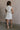 Full body back view of female model wearing the Jessica Off White Tortoise Button-Up Mini Dress which features Off White Fabric, Tortoise Button-Down Front Closure, Buttoned Cinch Details, Mini Length, Two Front Pockets, Two Front Chest Pockets, Sleeveless and Collared Neckline.