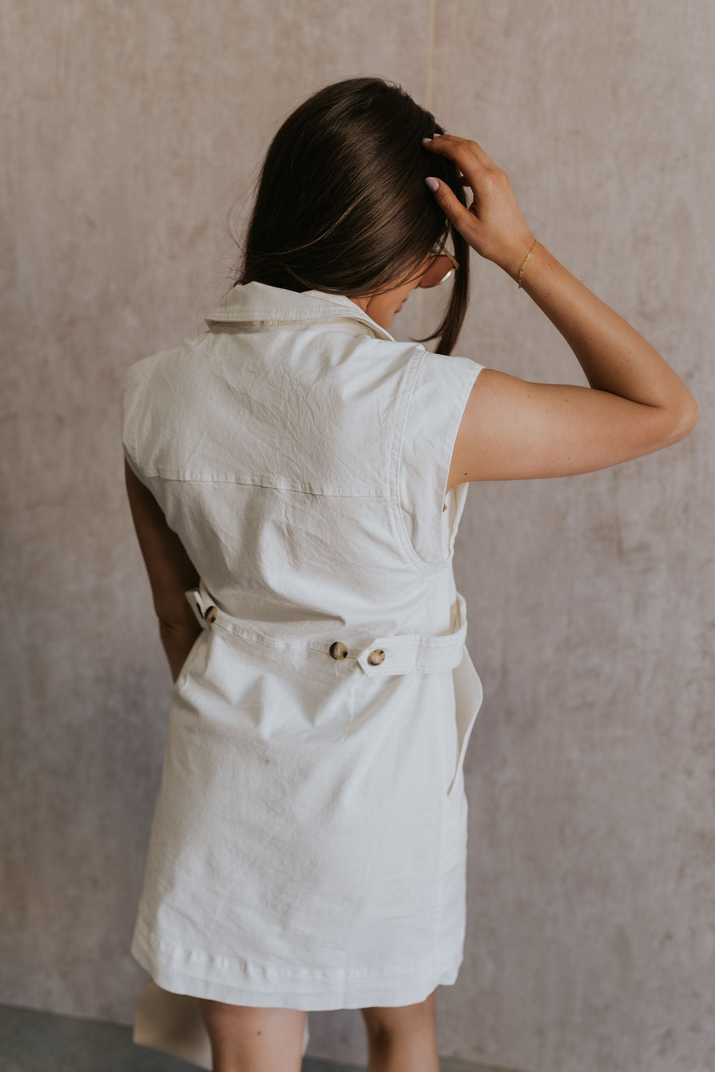 Back view of female model wearing the Jessica Off White Tortoise Button-Up Mini Dress which features Off White Fabric, Tortoise Button-Down Front Closure, Buttoned Cinch Details, Mini Length, Two Front Pockets, Two Front Chest Pockets, Sleeveless and Collared Neckline.