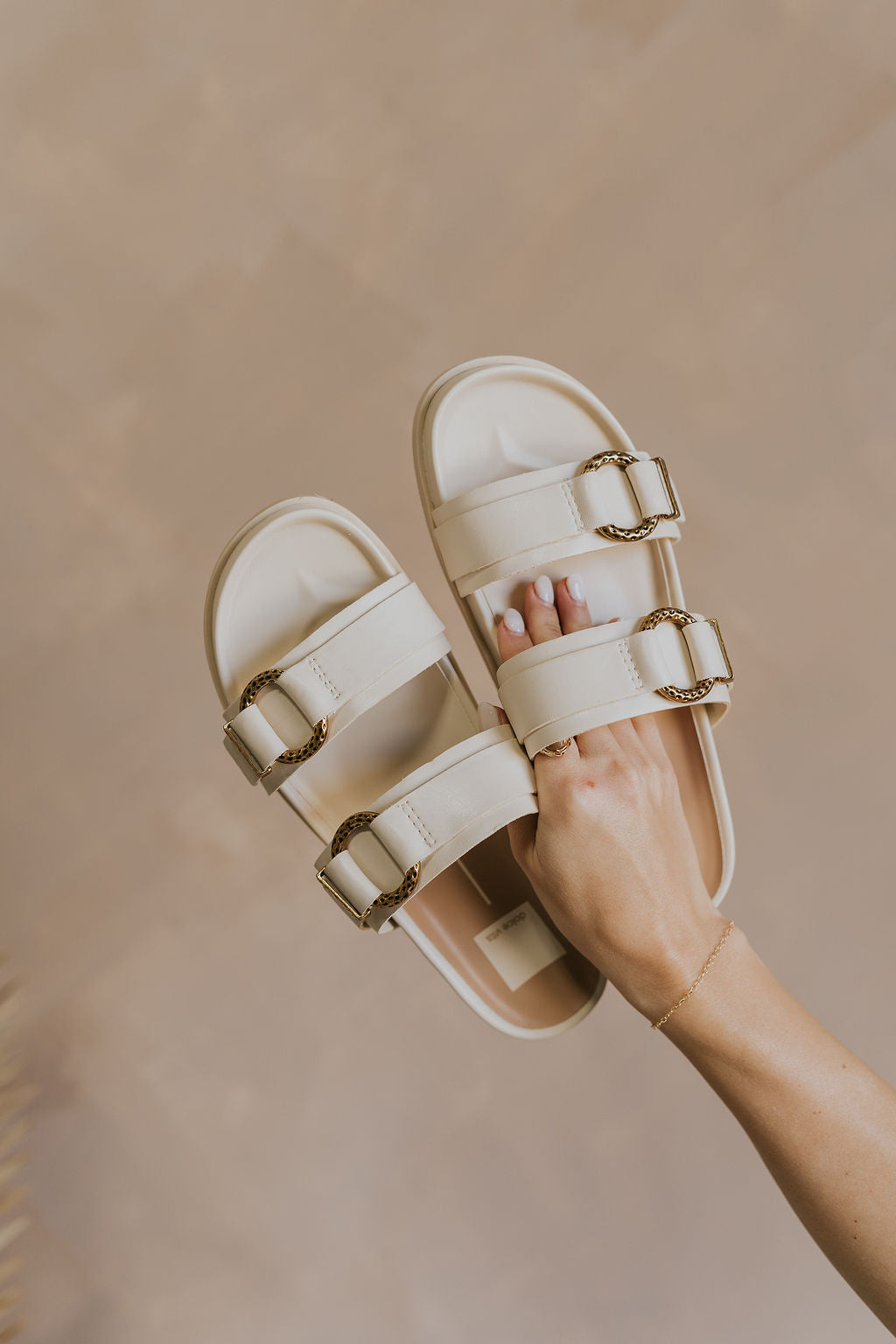 Front view of female model holding the Soya Sandal in White Leather which features white leather fabric, chunky sole, gold hammered metallic ring accents and slip-on style